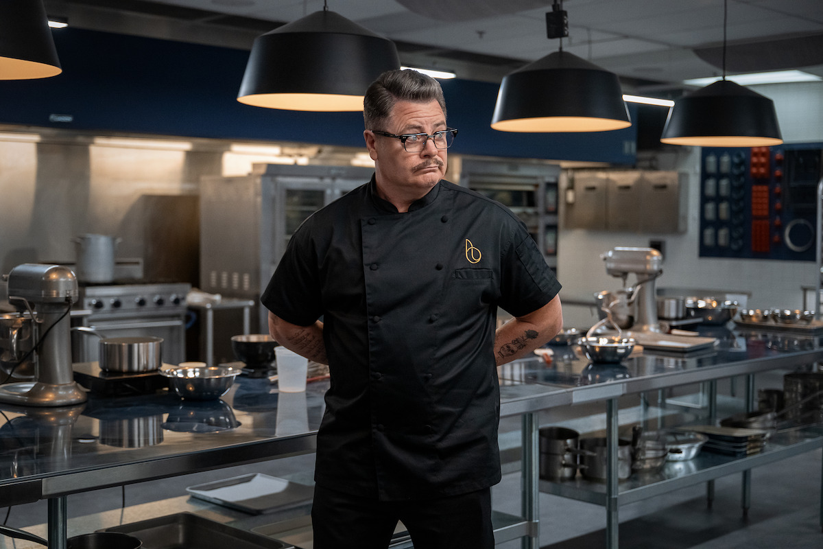 Male chef in black uniform standing in a modern, professional kitchen with stainless steel counters, mixers, and cookware under large black pendant lights.
