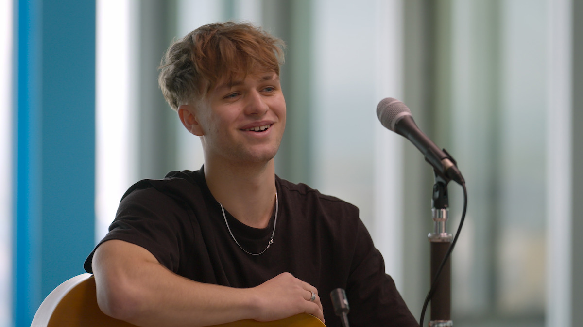 Young man with tousled hair smiles while sitting by a microphone holding a guitar in a modern, bright indoor space, creating a relaxed and cheerful atmosphere.