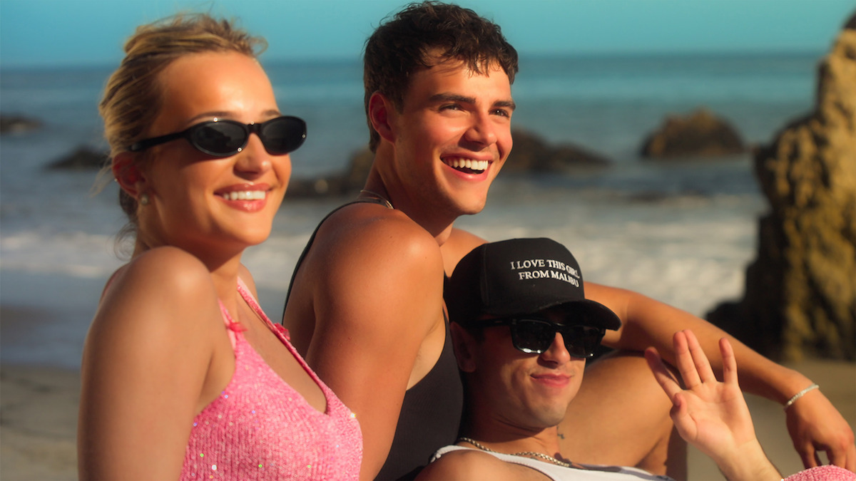 Three young adults relaxing on a sunny beach with rocks in the background, smiling and wearing sunglasses and casual beachwear.