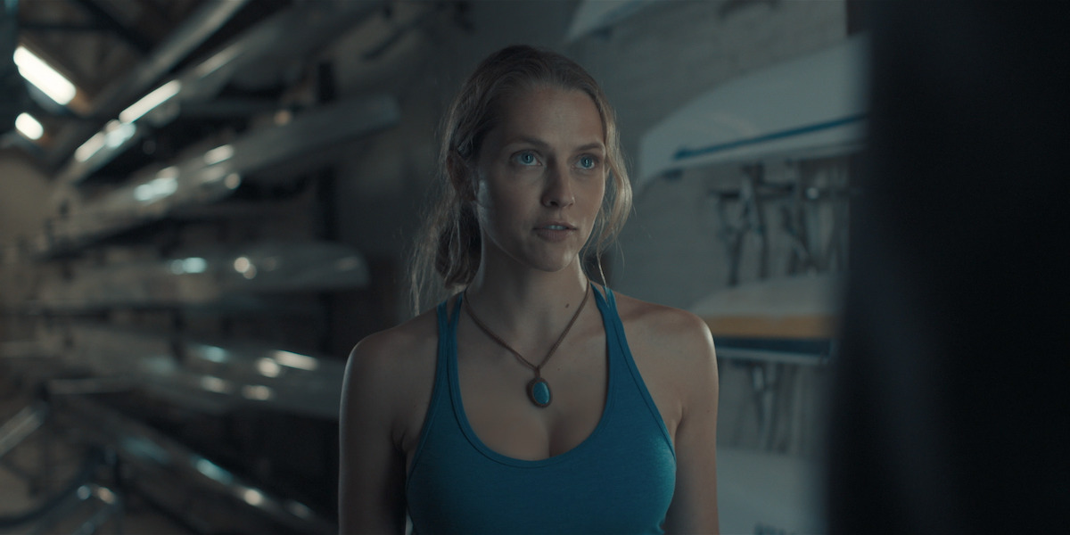 Teresa Palmer as Diana Bishop wears a blue tank top and stands in a corridor with canoes in an image from the series ‘A Discovery of Witches.’