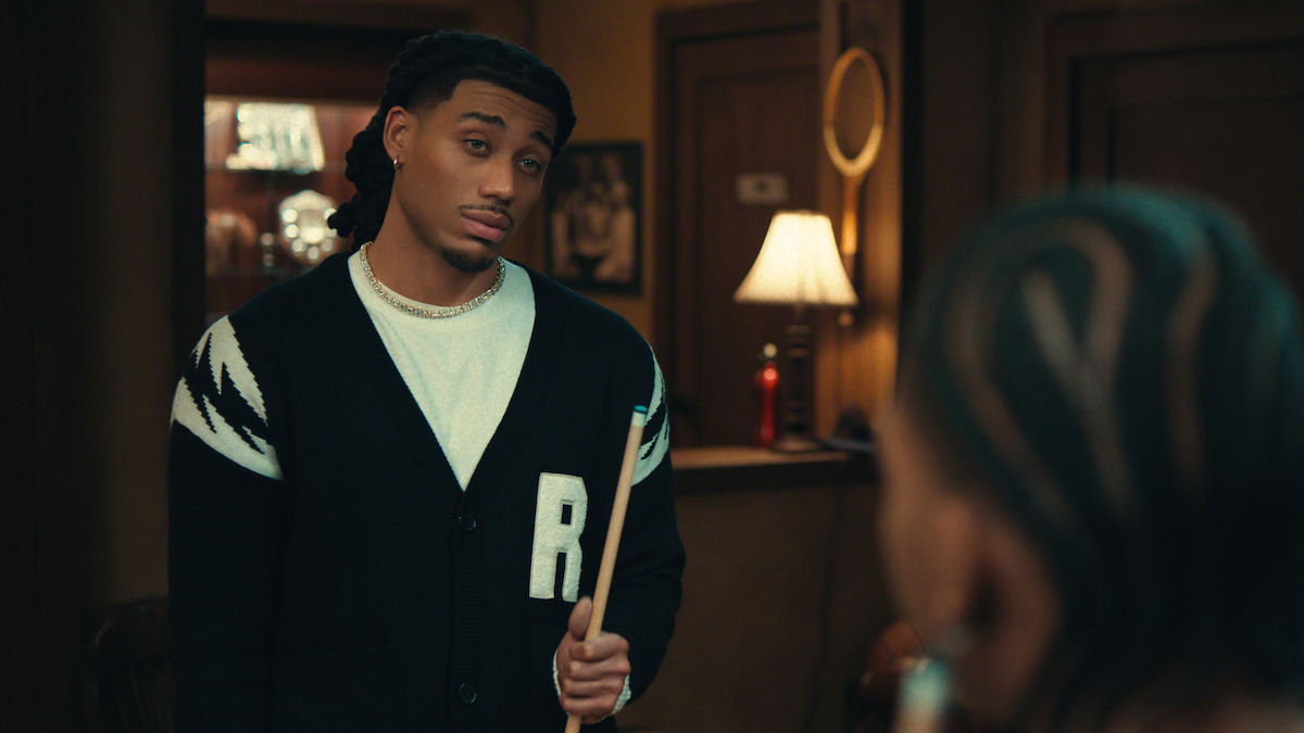 A man holding a pool cue stands in a warmly lit room, facing another person. There is a lamp on a table in the background, creating a cozy indoor environment with wood-paneled walls and decor.
