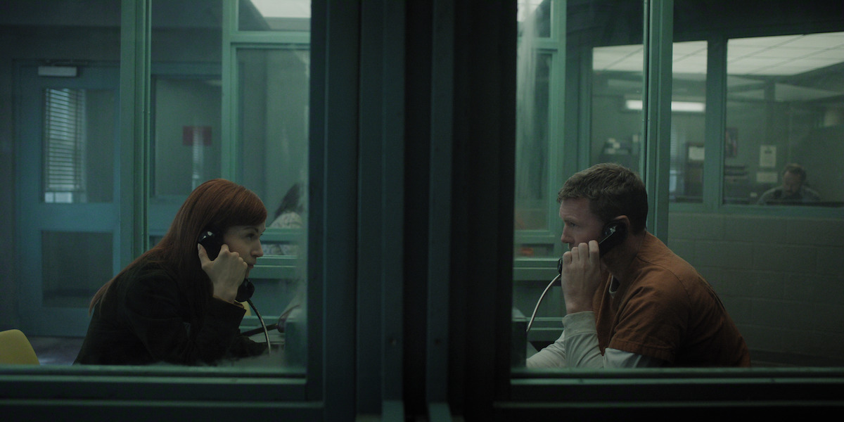 A woman and a man in prison uniform sit across glass in a visitation room using phones to talk. The setting is dimly lit with institutional green walls and other people are visible in the background.