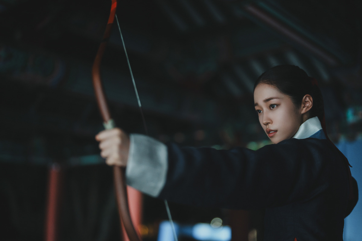 A young woman in traditional clothing focuses intently as she draws a bow in a dimly lit wooden structure, suggesting a historical or martial arts setting.