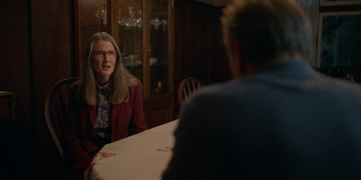 A woman with long gray hair and glasses sits at a dining table talking to a man in a dimly lit room with dark wood paneling and a glass cabinet in the background.