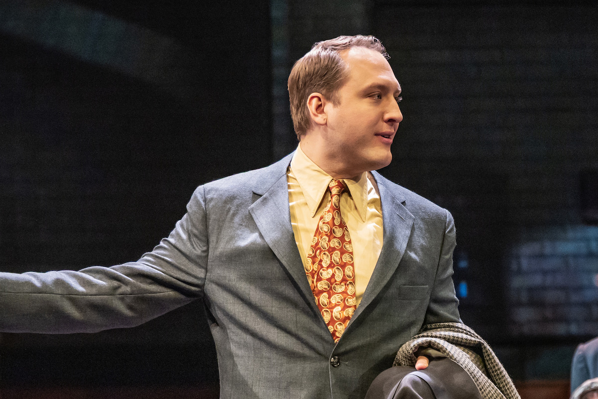 Man in a suit and patterned tie holding a coat and hat, standing on stage with a dark brick wall background, appearing to act or speak during a theater performance.