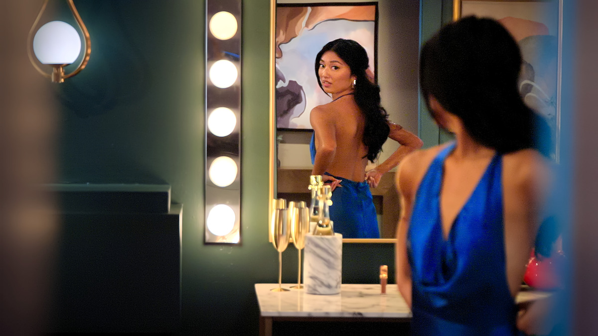 Emma Betsinger, in a blue dress, adjusts her outfit in front of a mirror in a stylish, dimly lit dressing room with wall lights, abstract art, and champagne glasses on a marble counter.