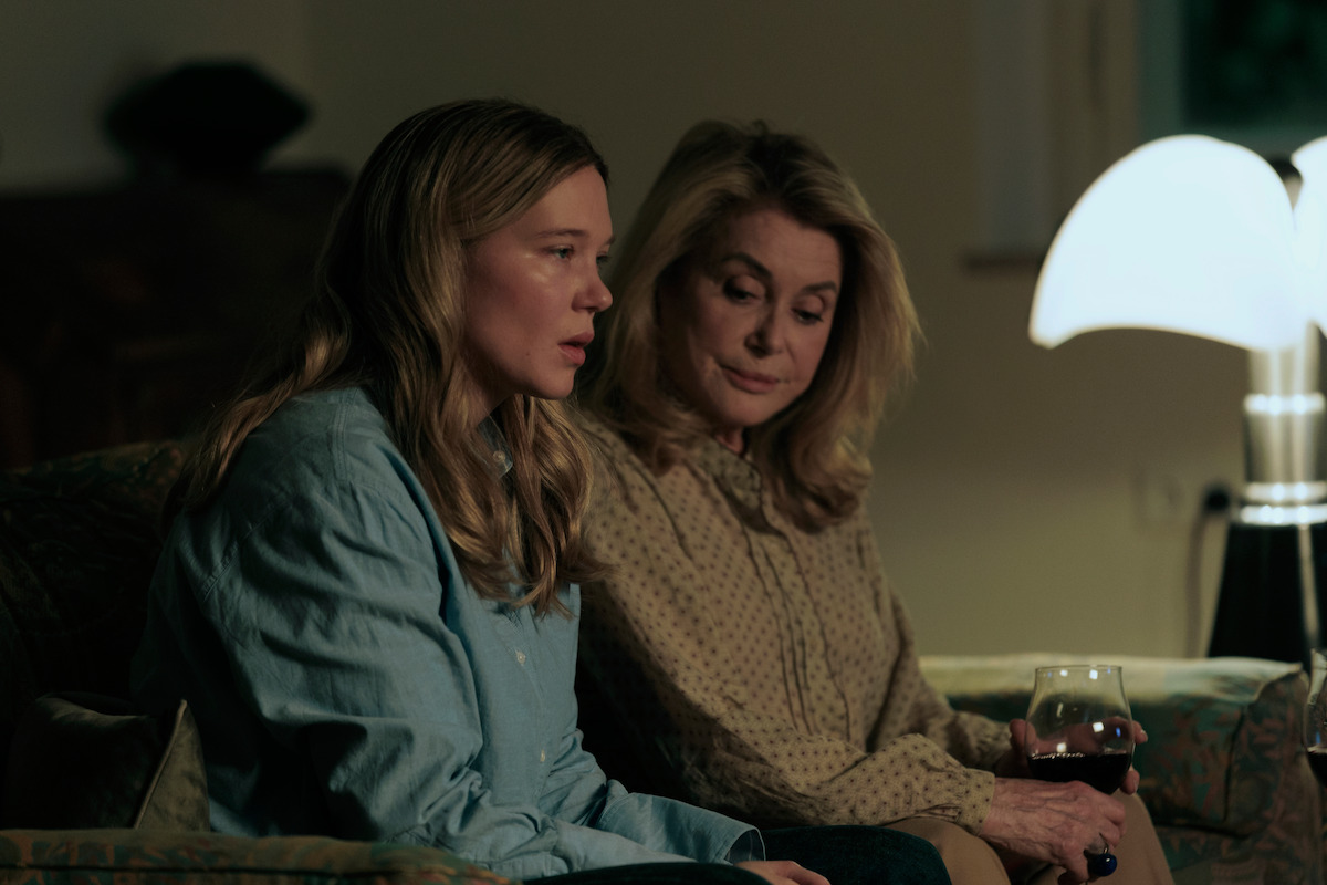 Two women sit on a couch in a dimly lit living room, one holding a glass of wine, both appearing thoughtful or serious, with a modern lamp providing soft light in the background.