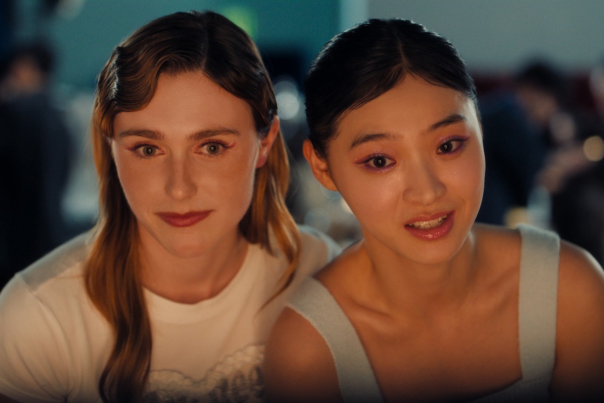 Two young women sit closely together indoors at an event, smiling softly at the camera with a blurred background suggesting a lively, social atmosphere.