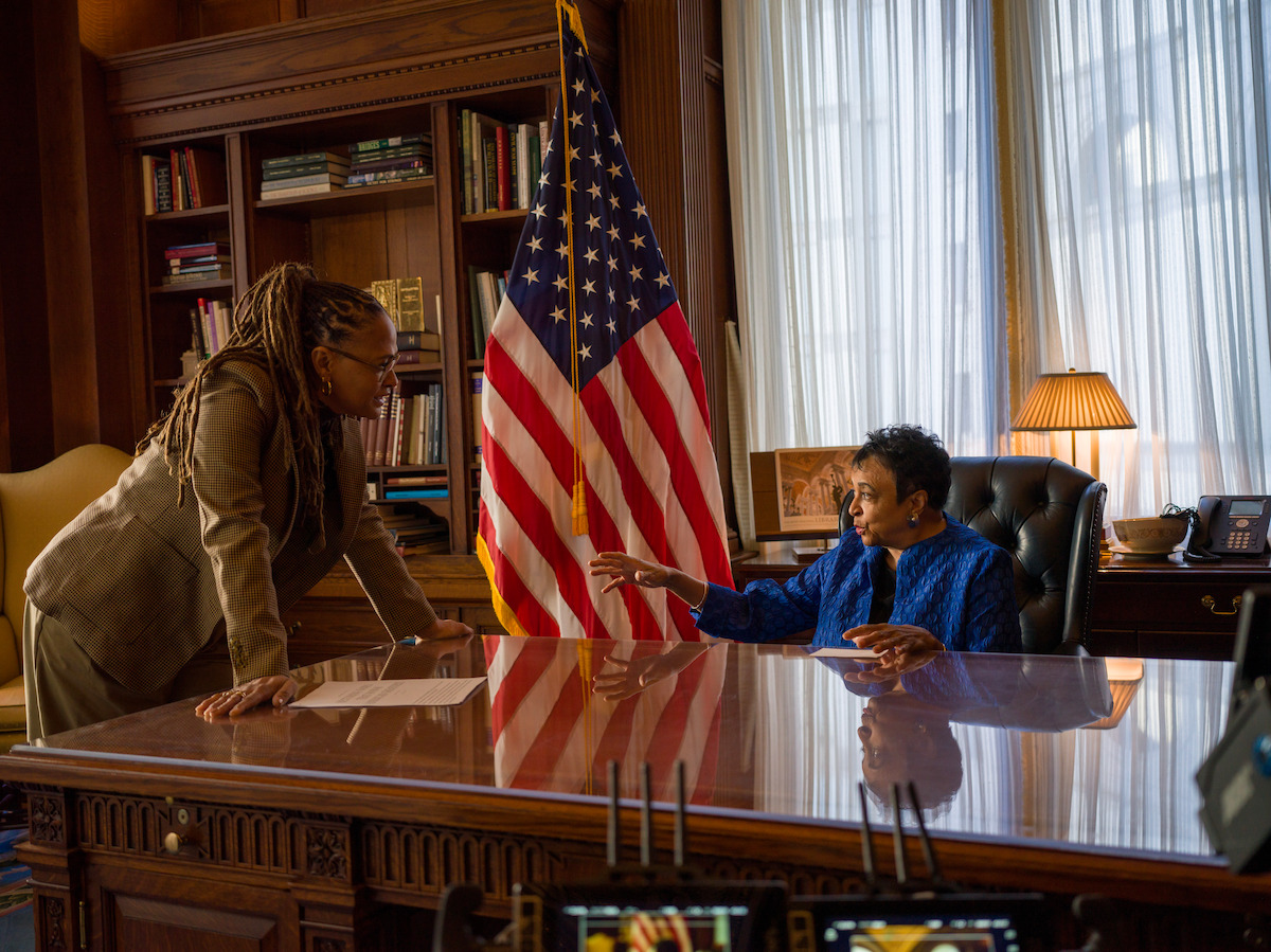 Two women converse in a formal office with a large wooden desk, bookshelves, and a US flag. One woman sits behind the desk gesturing, while the other stands leaning forward. Warm lighting and heavy drapery create an official atmosphere.