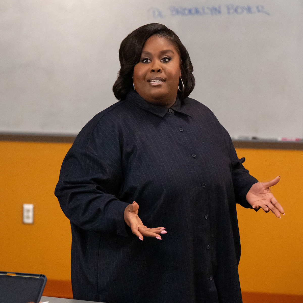 Raven Goodwin as Dr. Brooklyn Boyer speaking in a classroom, gesturing with hands, in front of a whiteboard with faint writing and orange walls in the background.