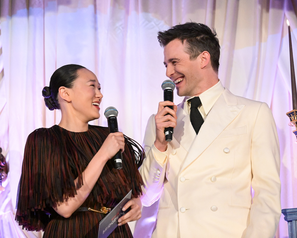 Two people smiling and holding microphones on stage, dressed formally in a dark dress and a white suit, standing in front of a light-colored curtain, sharing a joyful moment during an event.