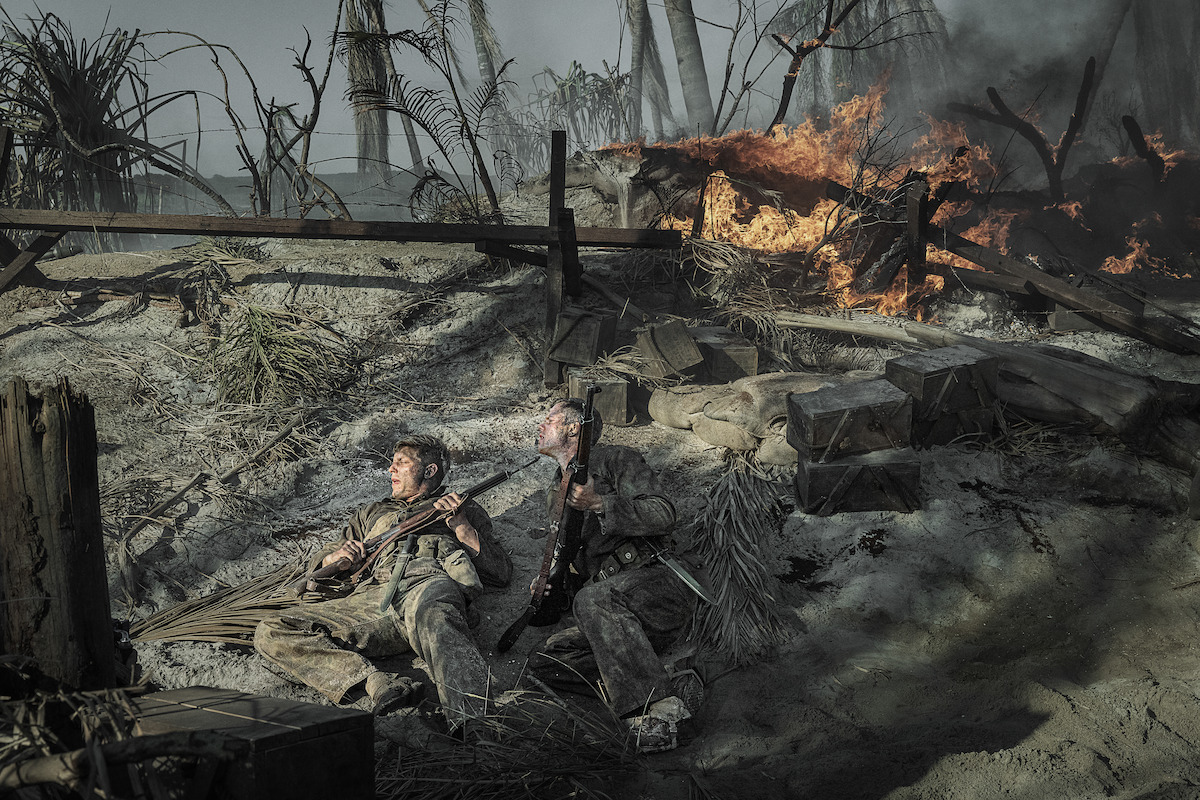 Two soldiers in dirty uniforms lie on a smoky, war-torn battlefield holding rifles, surrounded by debris and crates, as fire rages behind them and palm trees stand amid destruction.