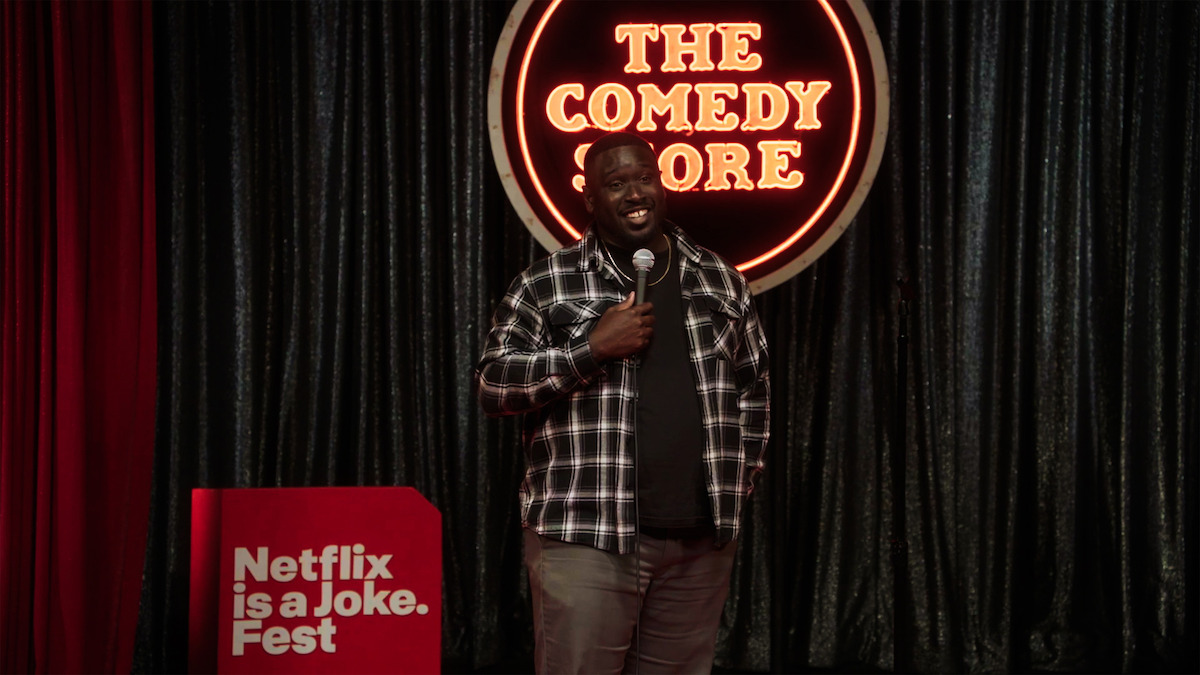 Comedian performing on stage at The Comedy Store during Netflix is a Joke Fest, holding a microphone in front of a black curtain with neon sign in the background.