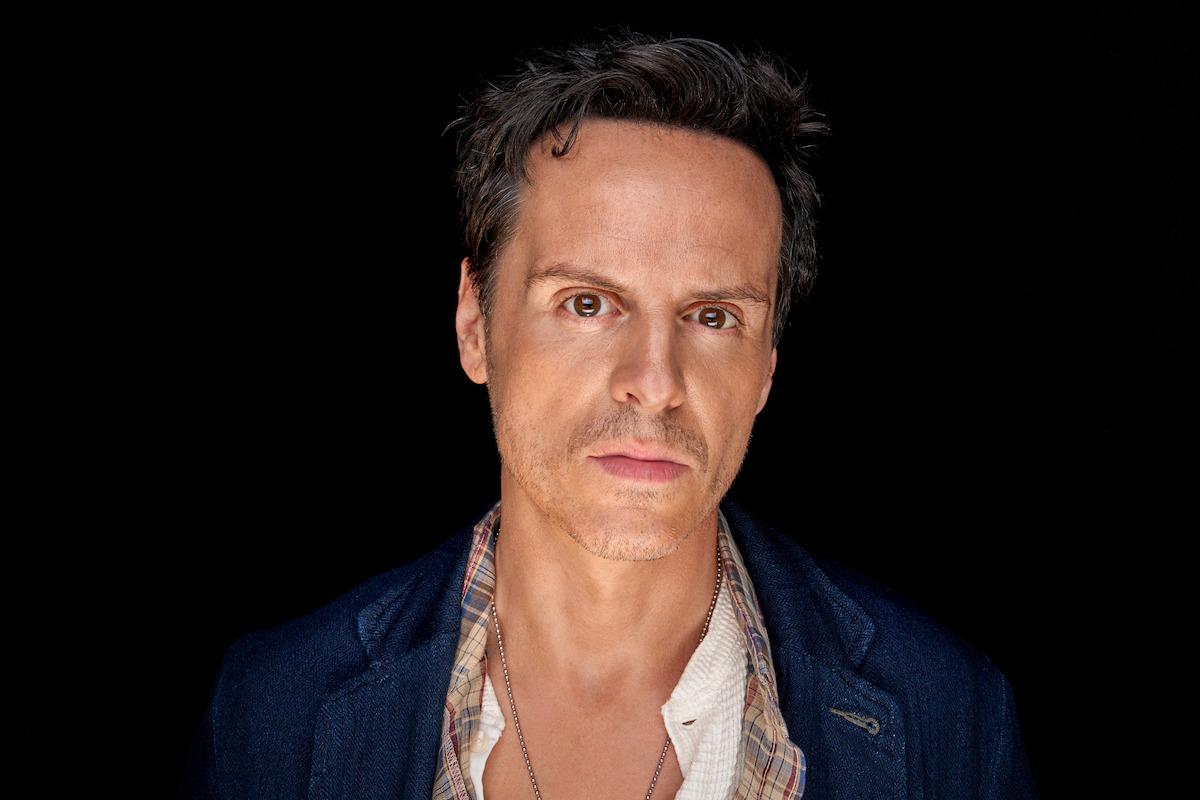 A man with short hair wearing a blue jacket and plaid shirt stands against a black background, looking directly at the camera with a serious and intense expression. Studio lighting creates a dramatic mood.