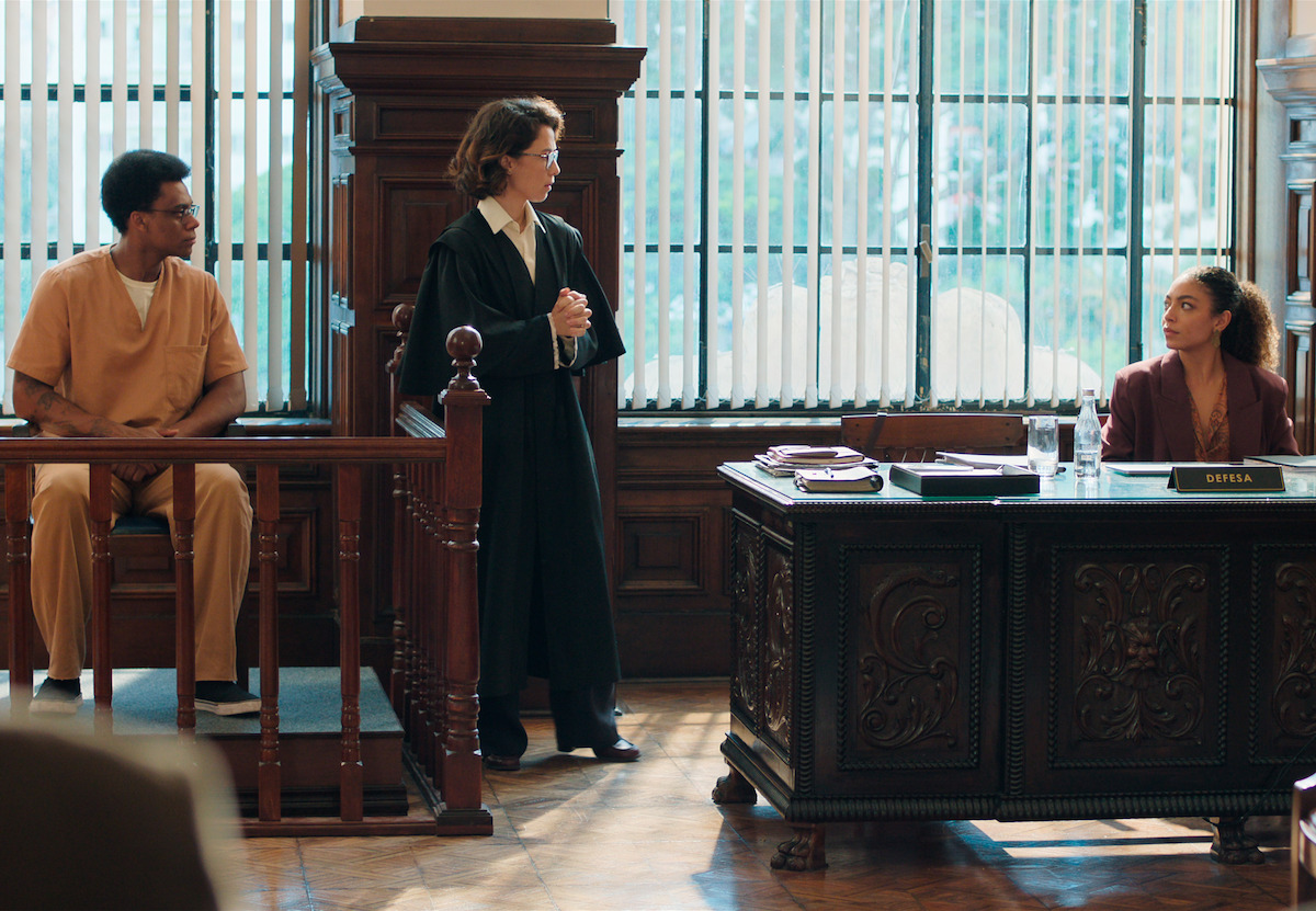 Em uma cena de tribunal, uma mulher (Marjorie Estiano) aparece em pé entre um homem, o réu, e sua advogada de defesa