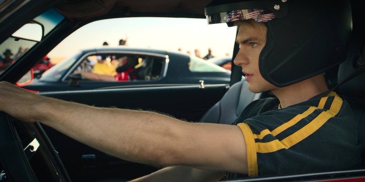 Young man wearing a racing helmet sitting in a car, gripping the steering wheel, preparing for a race. Other cars and people are visible in the background, suggesting a motorsport or racing event under daylight.
