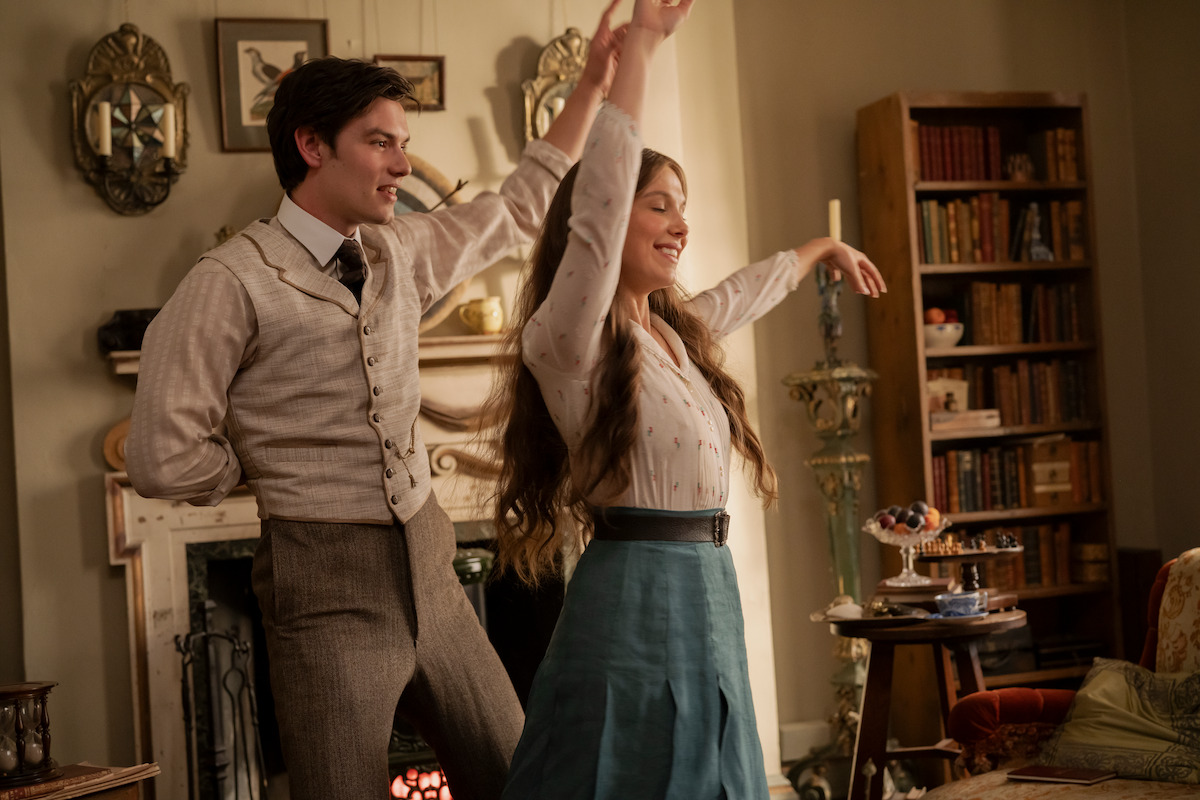 A man and woman in vintage clothing dance and smile in a cozy, well-furnished living room with bookshelves, a fireplace, and warm lighting, evoking a historical setting.