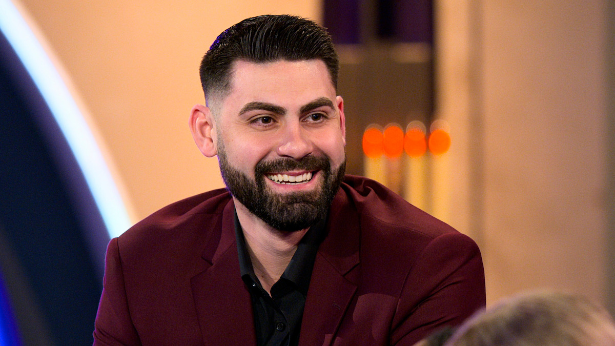 A bearded man in a burgundy jacket smiles while seated indoors on a brightly lit set, with blurred background elements and warm lighting.