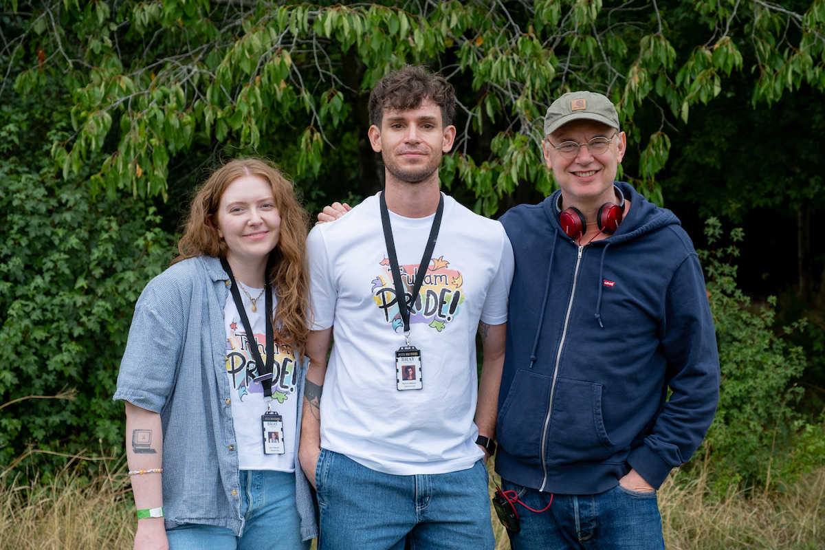 Three people smiling outdoors in front of trees, two wearing "Pride" shirts and lanyards, one with headphones and a cap, suggesting a casual event or festival in a natural setting.