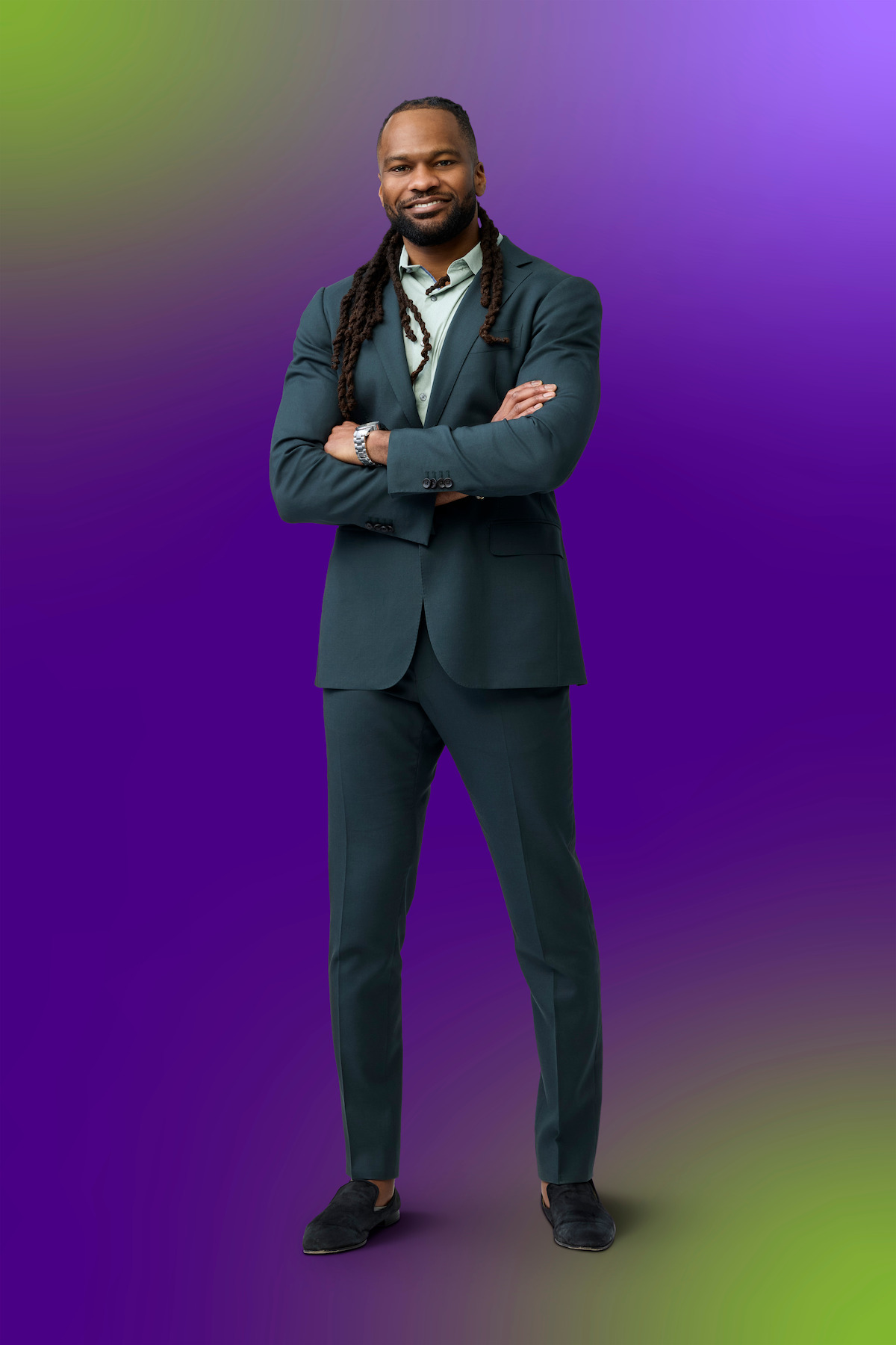 Man in a dark green suit standing confidently with arms crossed, smiling, on a purple and green gradient studio background.