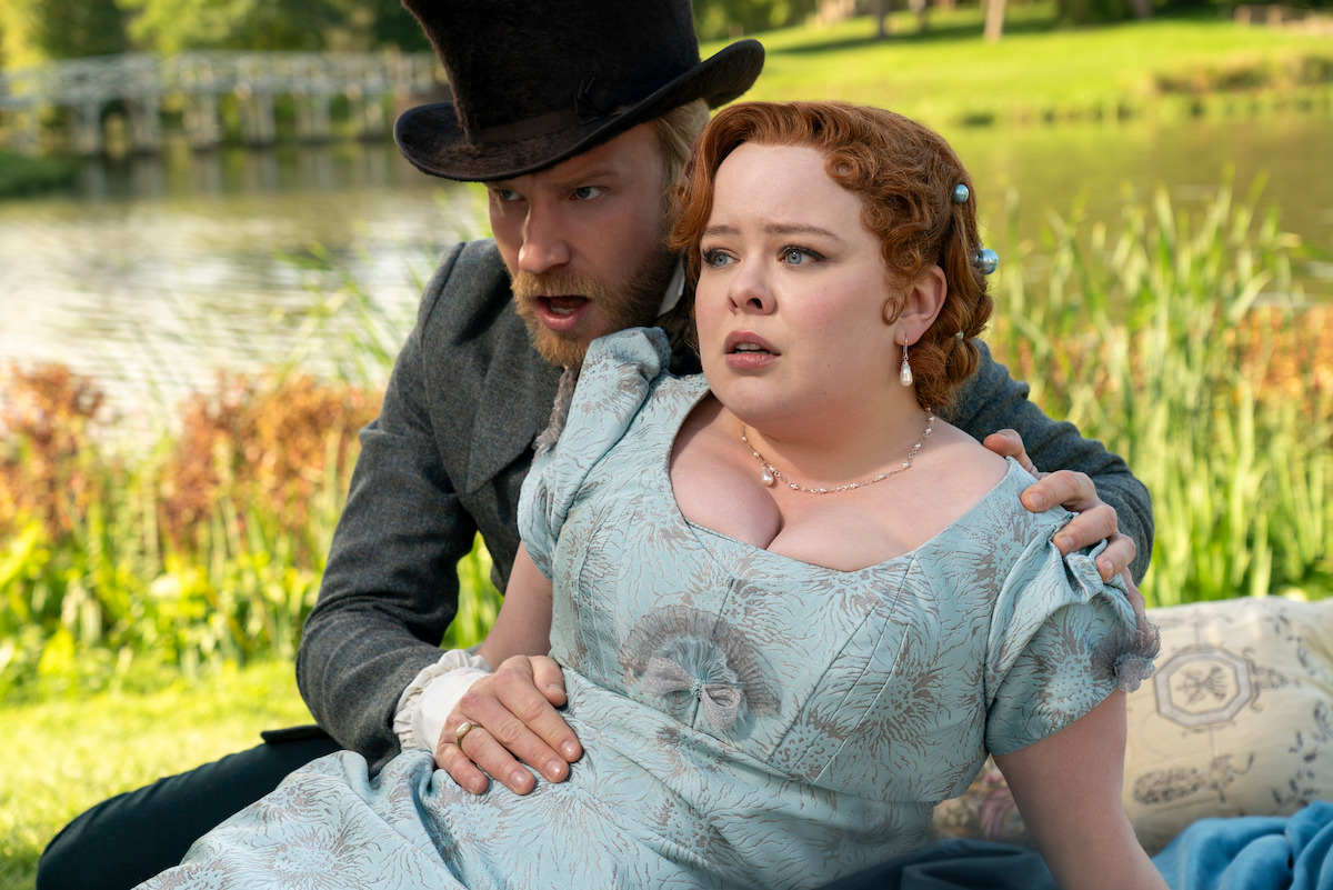 Sam Phillips as Lord Debling and Nicola Coughlan as Penelope Featherington sit on a grassy field with surprised expressions in Season 3 of ‘Bridgerton.’