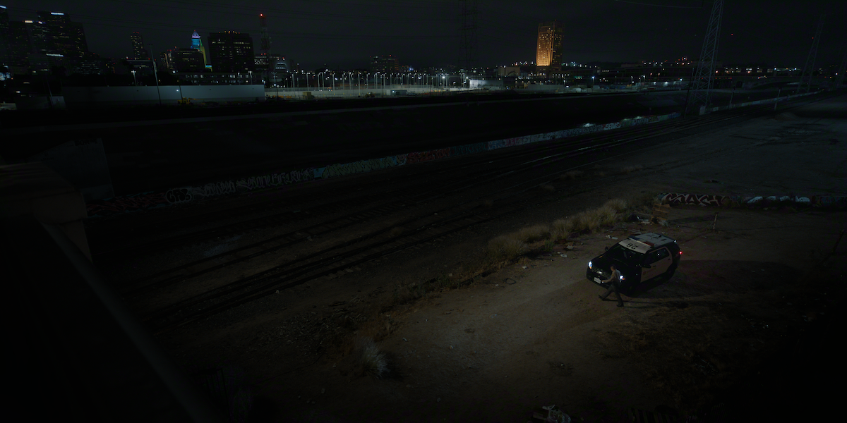 A police car parked on a dirt lot near railway tracks at night, surrounded by graffiti and dry brush, with a city skyline and illuminated buildings in the background.