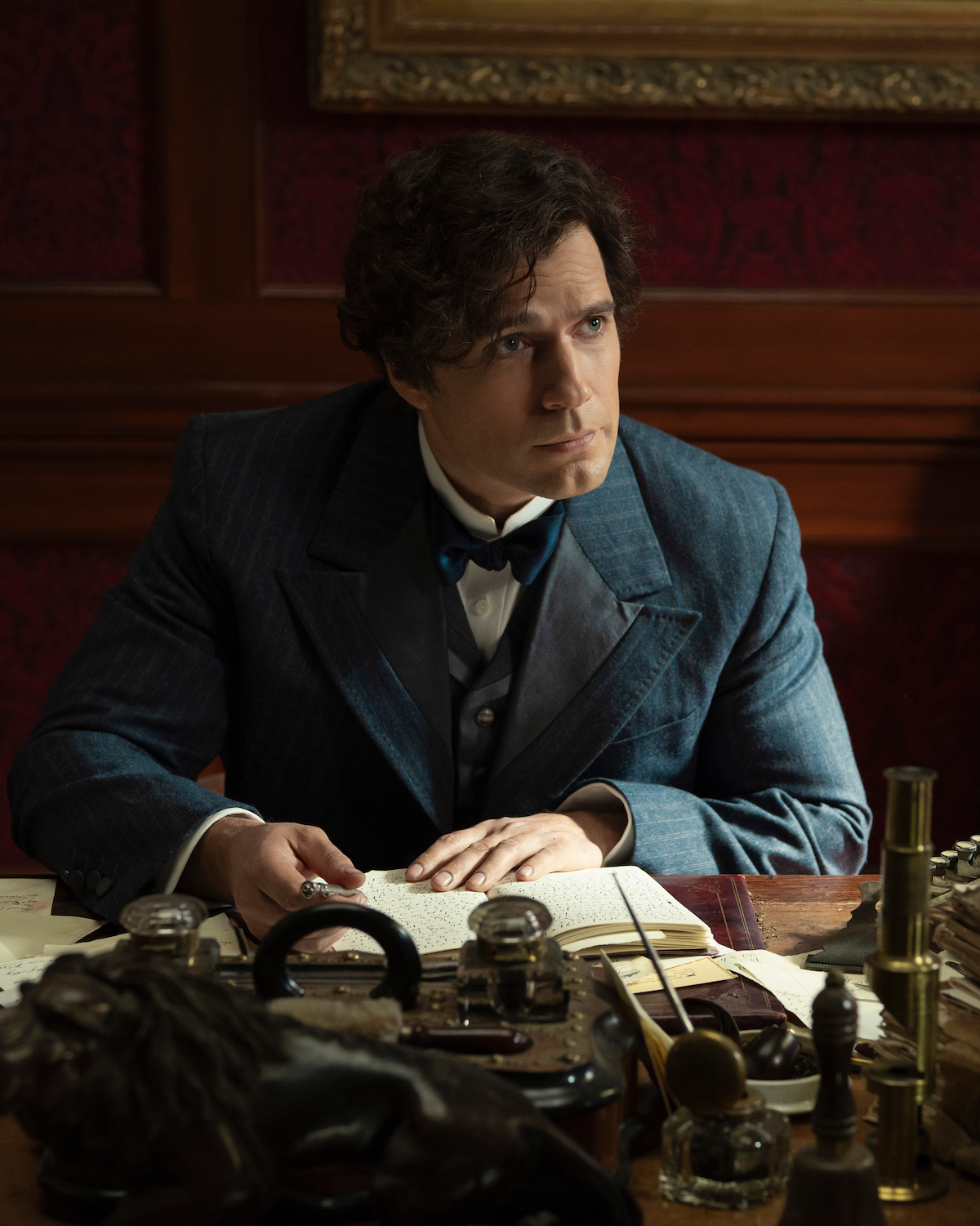 A man in vintage formal attire sits at a cluttered wooden desk with papers, books, and scientific instruments, in a richly decorated room with red wallpaper and framed artwork, appearing thoughtful or in the midst of writing.