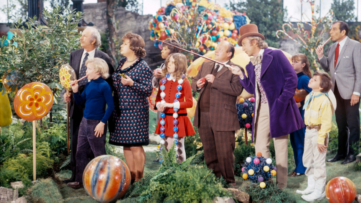 A group of people in colorful clothing stand in a whimsical candy-filled garden with oversized lollipops and sweets, surrounded by greenery and vibrant decorations, looking around in amazement.