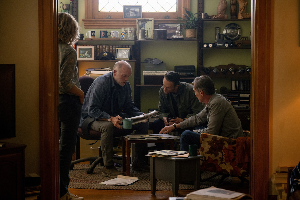 Four adults gathered in a cozy, warmly lit living room or study, seated around a coffee table covered in papers, books, and mugs, discussing or reviewing documents. Shelves with decor and plants in the background.