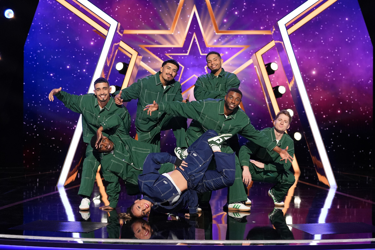 Rad Crew con chándales verdes posa en un escenario con un fondo morado estrellado y una estrella brillante, mientras un bailarín vestido de azul realiza un movimiento de breakdance en la pista de enfrente.