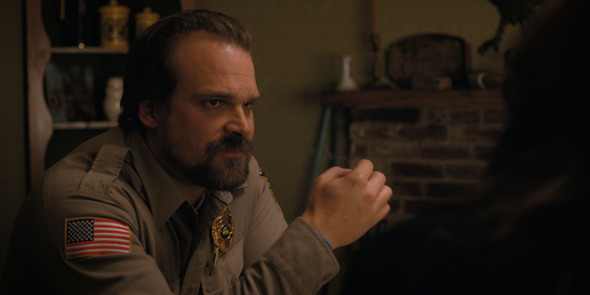 A serious police officer in uniform sits indoors at a table in a warmly lit room with rustic decor, a brick fireplace, and shelves, creating a tense and contemplative mood.