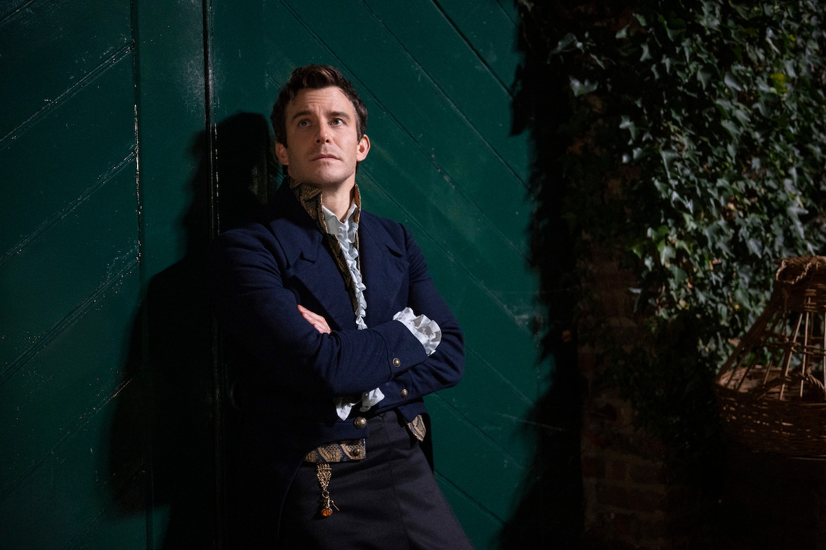 Man in period costume with arms crossed leans against a dark green wooden door, standing next to a brick wall covered in ivy, in a dimly lit outdoor setting.
