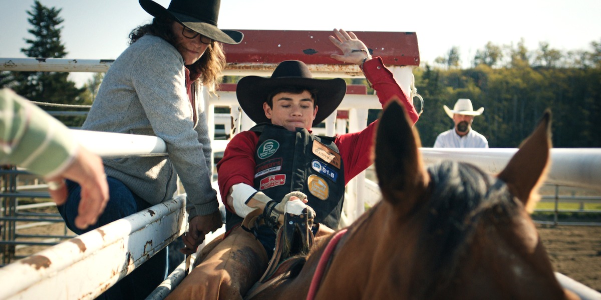 Young rodeo rider on horseback preparing for a ride in a rodeo arena, surrounded by people in cowboy hats and safety gear, outdoors, daytime.