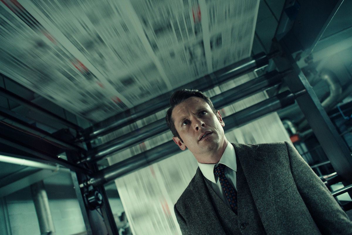 A worried man in a grey suit stands below a running newspaper printing press, pages blurring overhead.