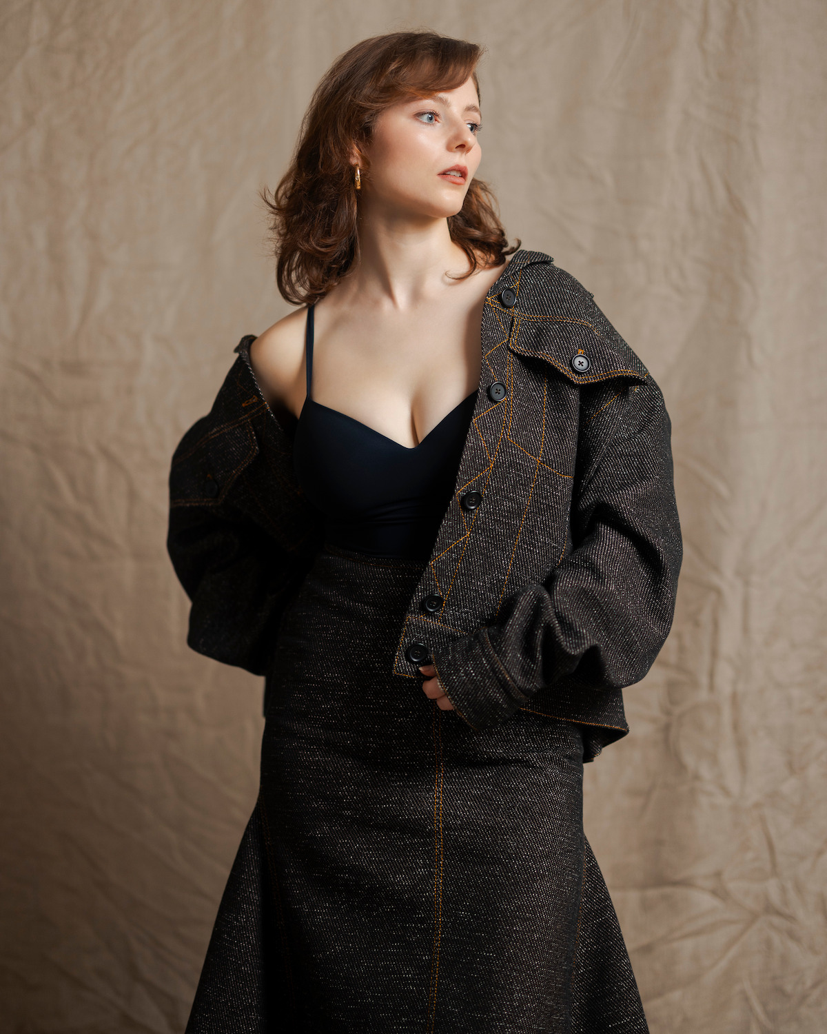 Thomasin McKenzie stands in front of a tan, wrinkled fabric backdrop wearing a dark denim jacket pulled down with one shoulder out and a matching denim skirt, looking over her shoulder.