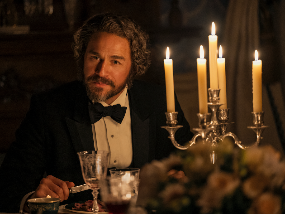 Man with curly hair and beard in formal attire sits at an elegant candlelit dinner table, surrounded by glasses, dishes, and a floral centerpiece in a warmly lit, upscale setting.