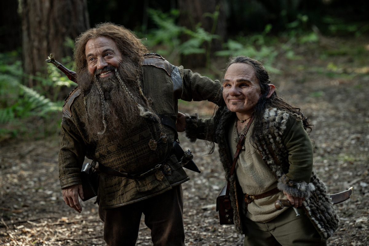 Two fantasy characters with medieval costumes and weapons stand in a lush forest, smiling and appearing cheerful, suggesting an adventurous and friendly mood in a natural, wooded setting.