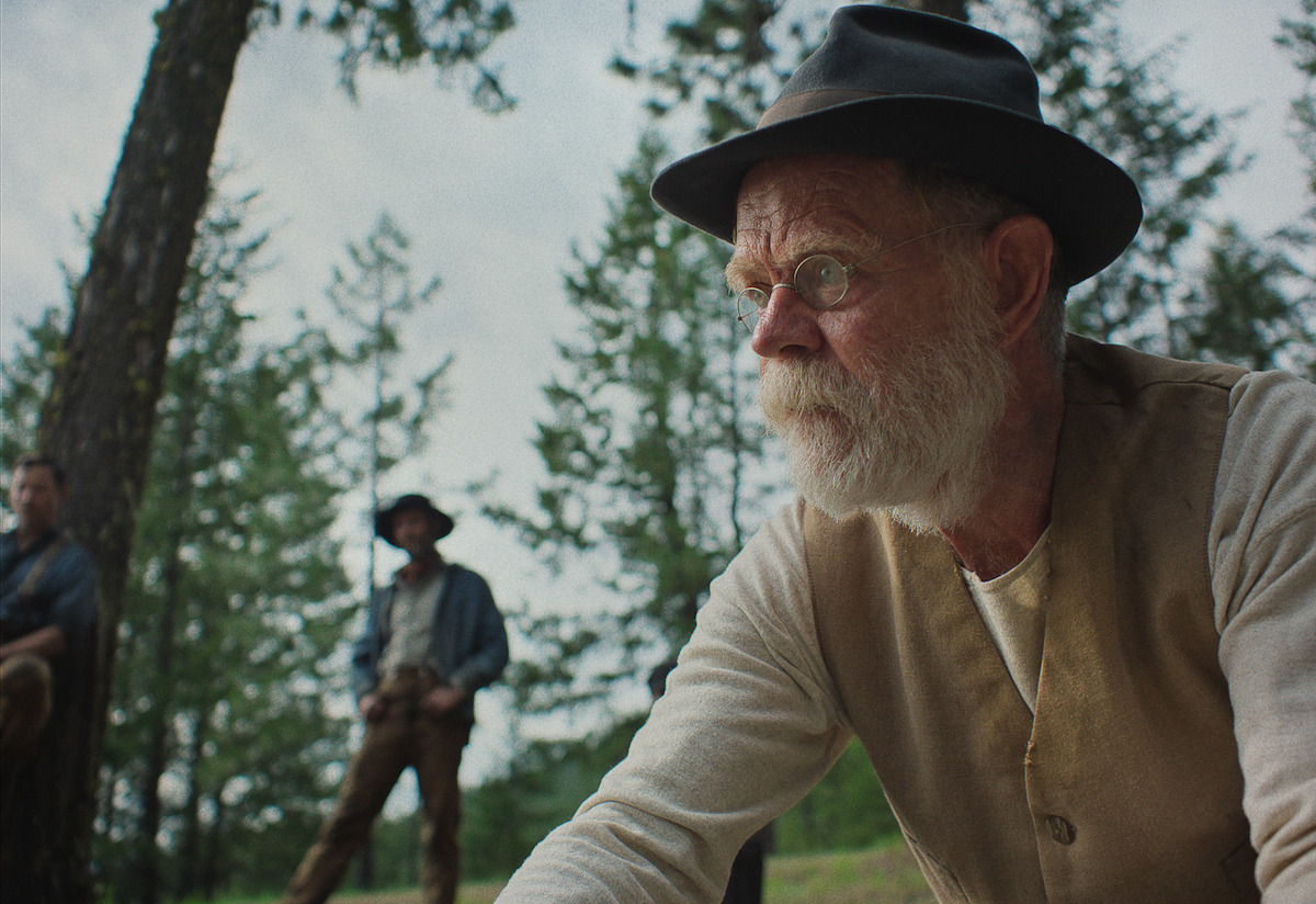 Elderly man in a hat and vest looks serious in a forest, with two other men standing in the background; the mood is tense and contemplative.