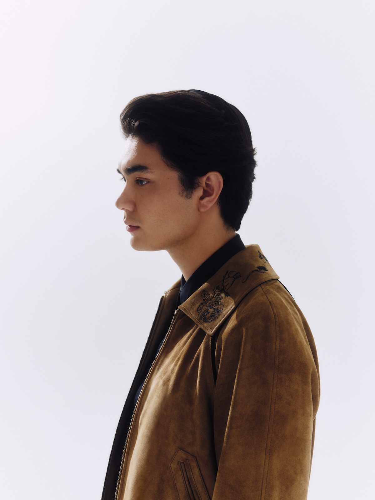 Young man in profile, wearing a brown suede jacket with embroidery, standing against a plain white background, soft studio lighting highlighting his features.