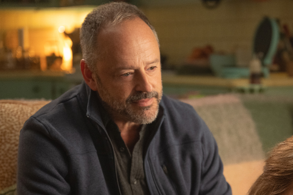 Middle-aged man with a beard sitting indoors on a couch, wearing a dark jacket, with a thoughtful expression in a warmly lit, cozy kitchen environment.