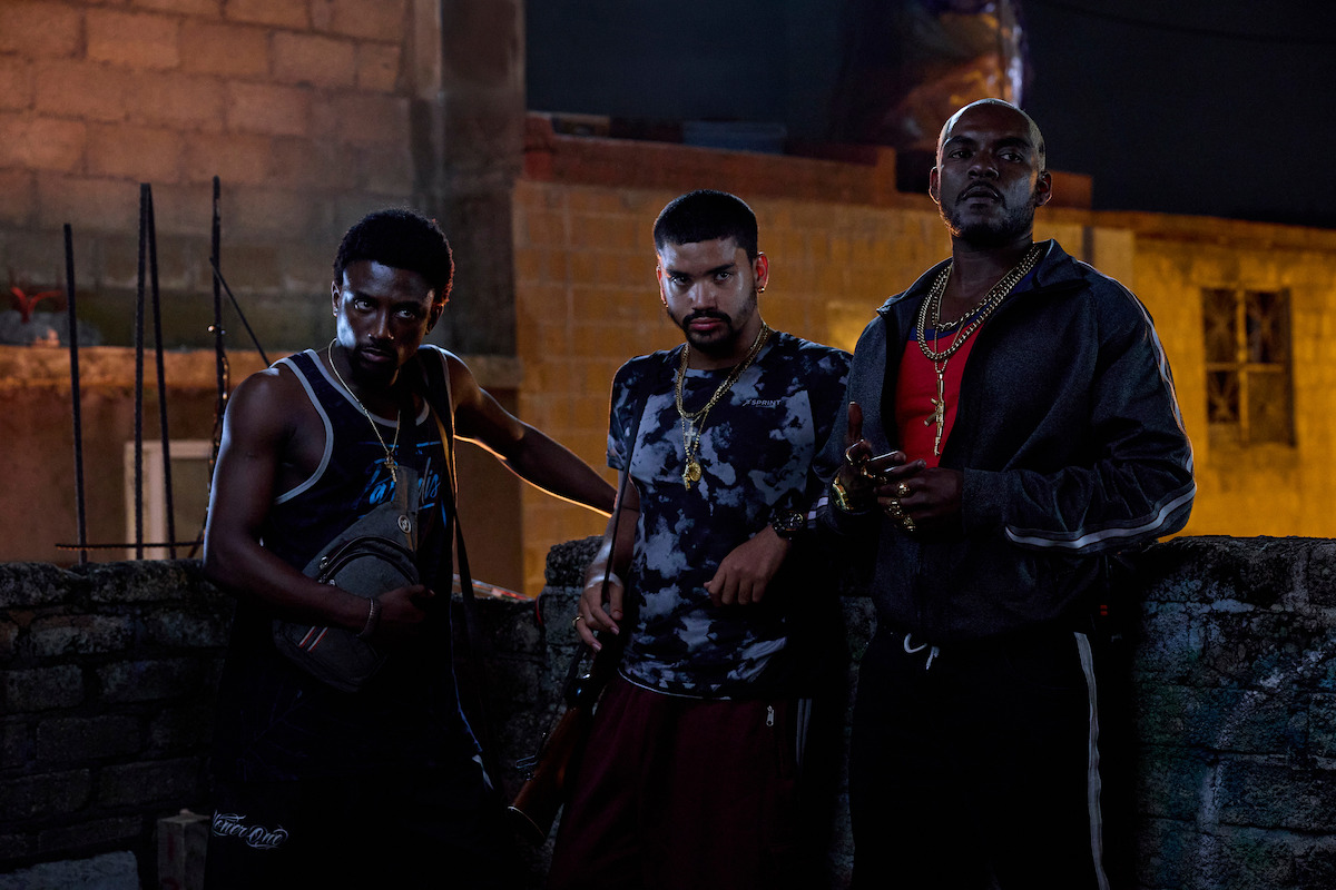 Three men with serious expressions stand behind a rough brick wall at night in an urban outdoor setting, wearing casual streetwear and jewelry, dimly lit by artificial lighting.