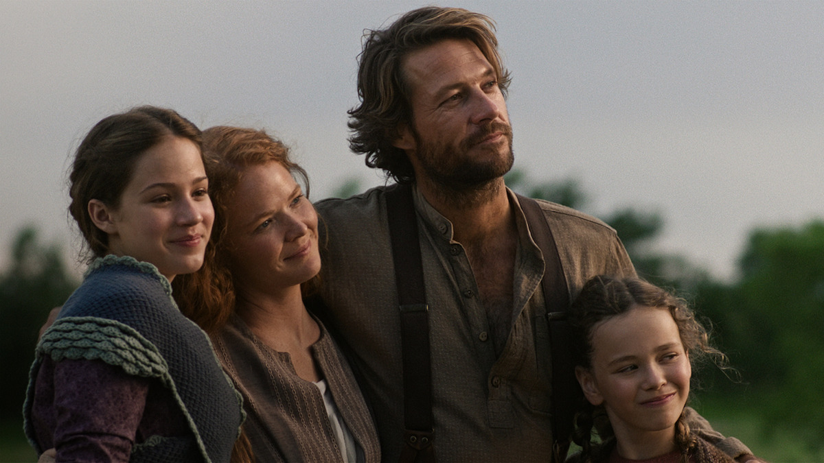 A family of four stands together outdoors, dressed in period clothing, smiling and embracing each other with greenery and a cloudy sky in the background.