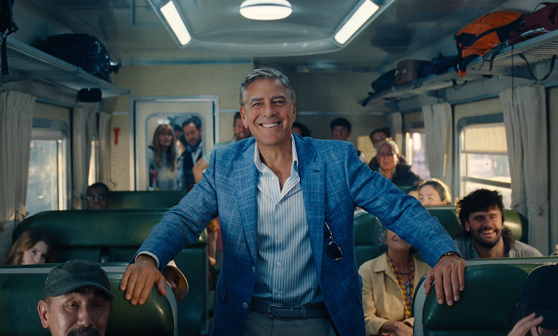 Jay Kelly (George Clooney) wears a suit and stands amongst the civilians on board of the passenger train in Italy with a big smile on his face.