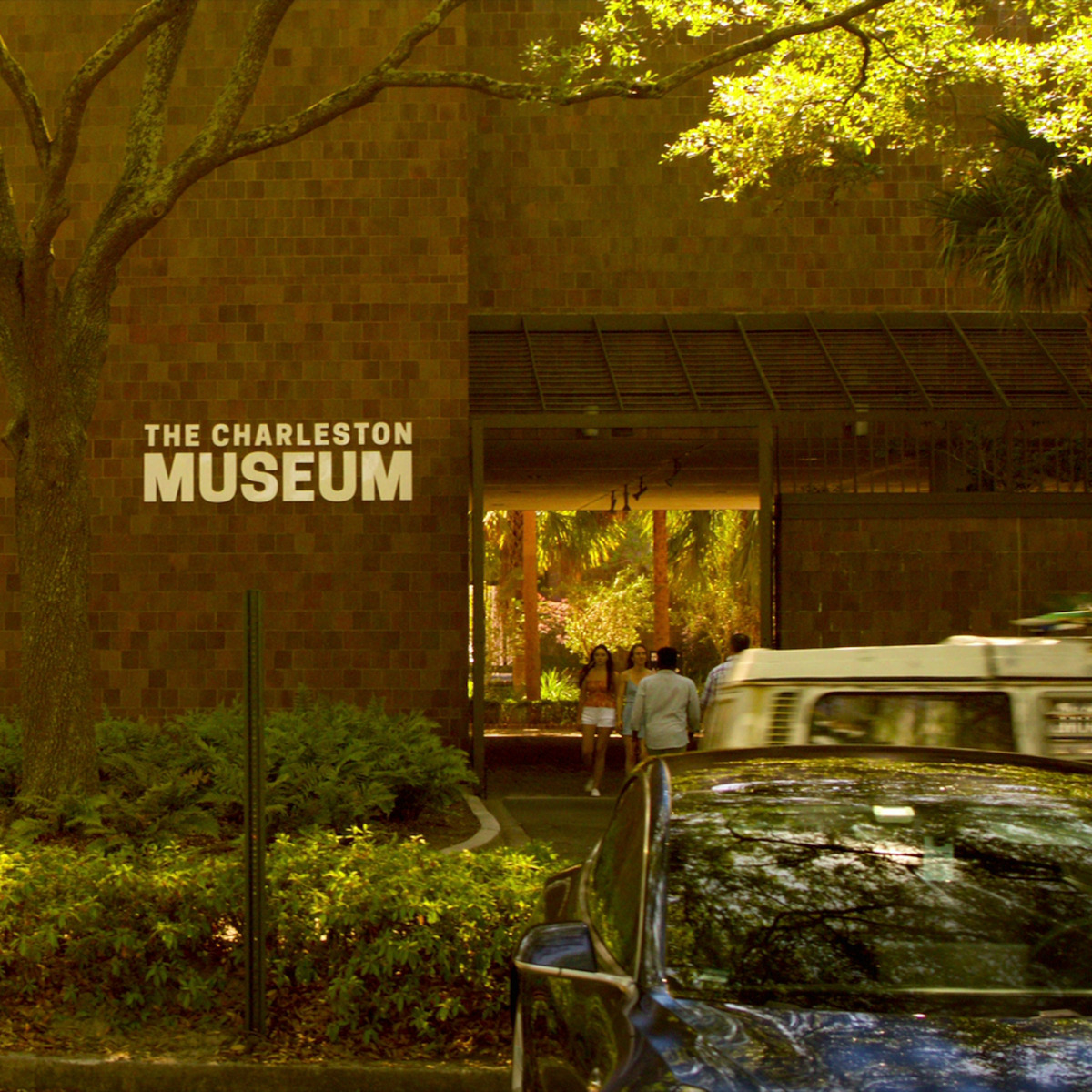 Entrada do Museu de Charleston, cercada de árvores e paisagem verde, com pessoas andando e carros estacionados na frente do imóvel em um dia de sol.