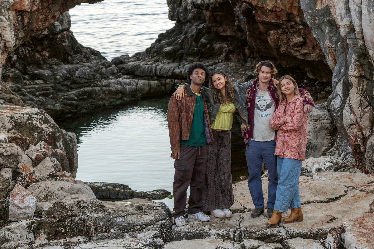 Four friends stand together smiling on rocky terrain near a natural pool and ocean, framed by rock formations, suggesting a relaxed and adventurous outdoor setting.