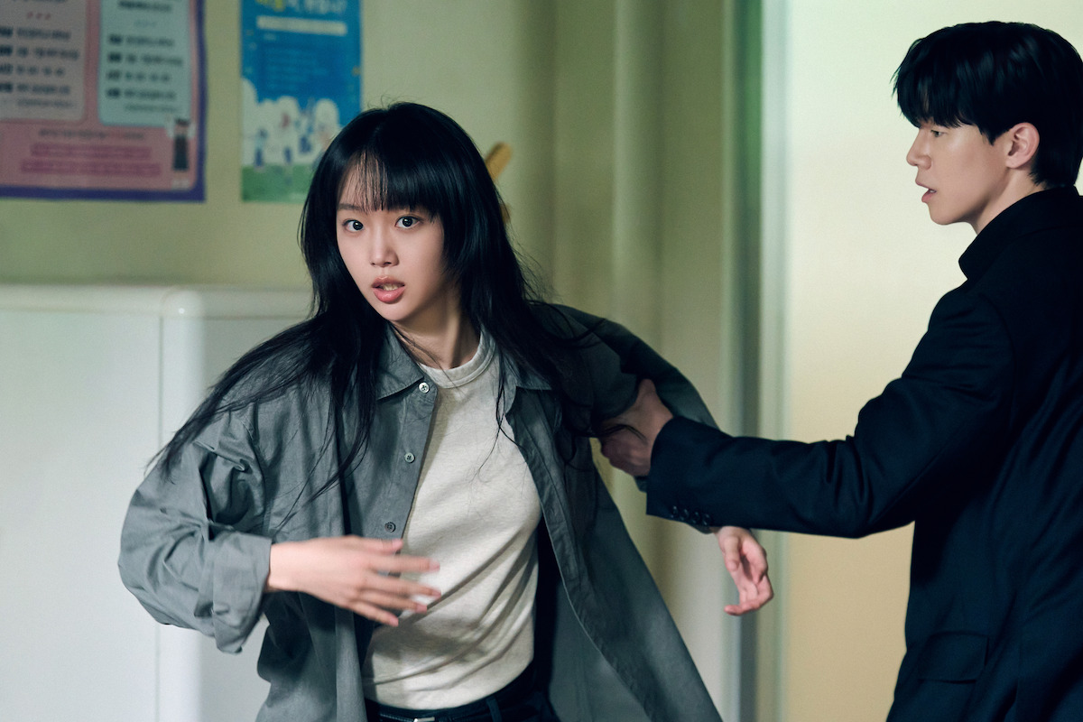A woman in green jacket looks off into the distance while a man grabs her by the arm as she attempts to break away.