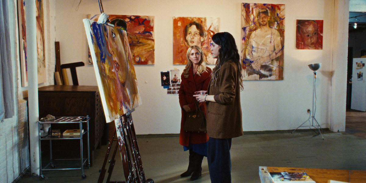 Two women in an art studio discuss a large, colorful portrait on an easel, surrounded by expressive paintings on the wall. The setting is creative and intimate, suggesting a focused, artistic mood.