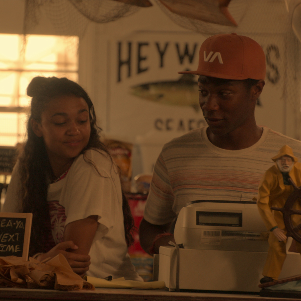Duas pessoas jovens estão em pé atrás do balcão em um restaurante de frutos do mar, com redes e uma placa onde se lê "Heywards Seafood" ao fundo, uma caixa registradora o boneco de um pescador amarelo no balcão, com iluminação aconchegante.