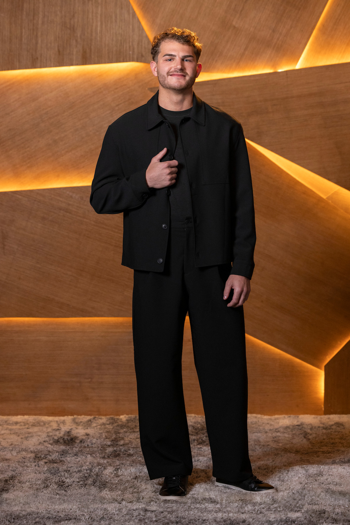 A man in a black outfit stands on a carpeted floor in front of a modern wood-paneled wall with glowing yellow light accents, posing and smiling at the camera.
