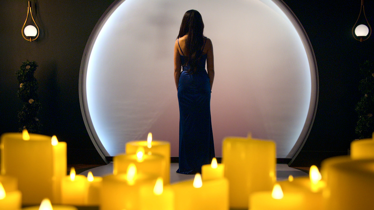 Woman in a blue dress stands facing a glowing circular backdrop, surrounded by lit yellow candles in a dimly lit, elegant room with plants and modern wall sconces.